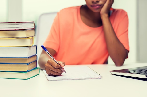 koncepcja ludzi, technologii, nauki i edukacji - zbliżenie znudzonej Afroamerykanki młodej kobiety siedzącej przy stole z laptopem i książkami oraz piszącej do notebooka w domu