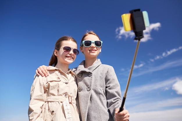 koncepcja ludzie, dzieci, przyjaciele i przyjaciele - szczęśliwe dziewczyny robiące zdjęcie ze smartfonem na kij do selfie na zewnątrz