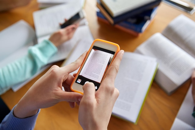 koncepcja ludzie, edukacja, technologia i egzamin - zbliżenie uczniów ze smartfonami robiących zdjęcie strony książek i robiących ściągawkę w szkolnej bibliotece