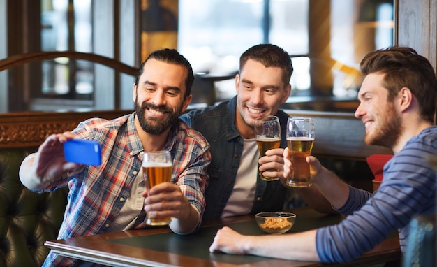 koncepcja ludzie, mężczyźni, wypoczynek, przyjaźń i technologia - szczęśliwi mężczyźni pijący piwo i robiący selfie ze smartfonem w barze lub pubie