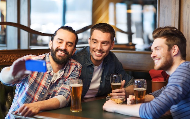koncepcja ludzie, mężczyźni, wypoczynek, przyjaźń i technologia - szczęśliwi mężczyźni pijący piwo i robiący selfie ze smartfonem w barze lub pubie
