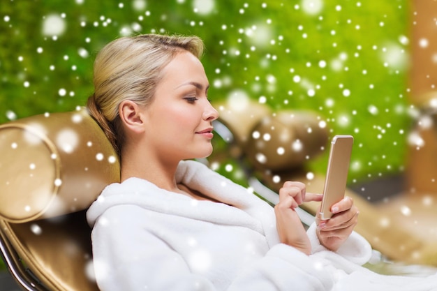 koncepcja ludzie, piękno, styl życia, technologia i relaks - piękna młoda kobieta w białym szlafroku ze smartfonem w sieci społecznościowej w spa z efektem śniegu