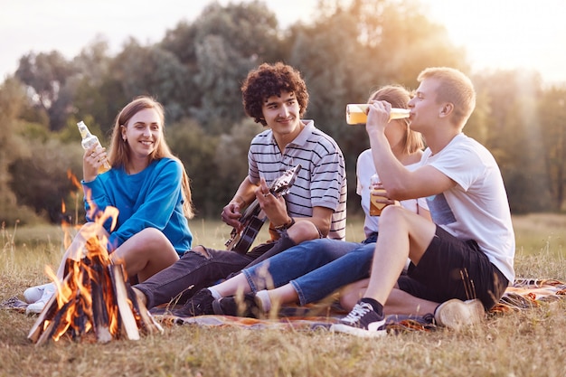 Koncepcja Ludzie, Piknik I Styl życia. Radośni Czterej Przyjaciele śpiewają Piosenki I Grają Na Gitarze, Siedzą Przy Ognisku, Cieszą Się Letnim Drinkiem, Oddychają świeżym Powietrzem