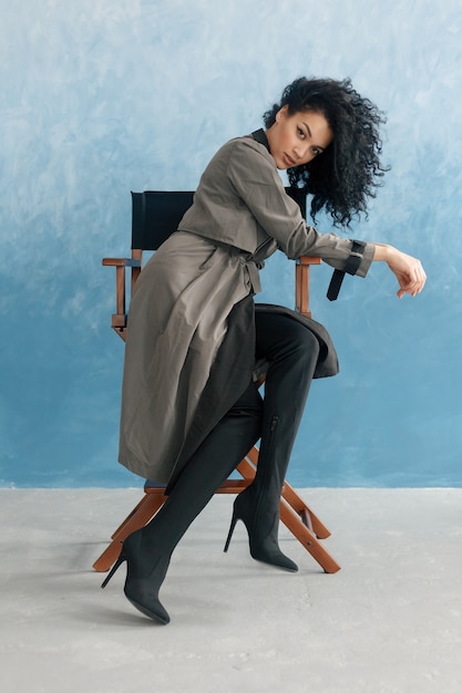 Koncepcja ludzie, rasa, pochodzenie etniczne i moda - uroda czarny model pozowanie w wysokich butach. Młoda piękna kobieta Mulat siedzi na krześle reżysera w filmie. kobieta w szarym płaszczu przeciwdeszczowym, fryzura afro