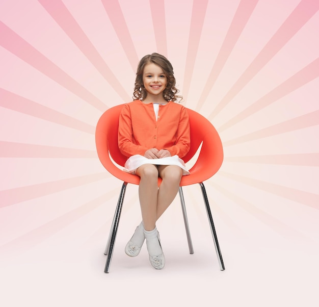 koncepcja ludzie, szczęście, dzieciństwo i meble - szczęśliwa mała dziewczynka siedzi na designerskim krześle na różowym tle promieni wybuchających