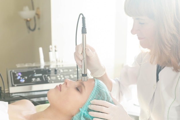 Koncepcja Ludzie, Uroda, Spa, Kosmetologia I Technologia - Kosmetolog Wykonuje Zabieg Mikroprądowej Terapii Skóry Twarzy Pięknej, Młodej Kobiety W Gabinecie Kosmetycznym