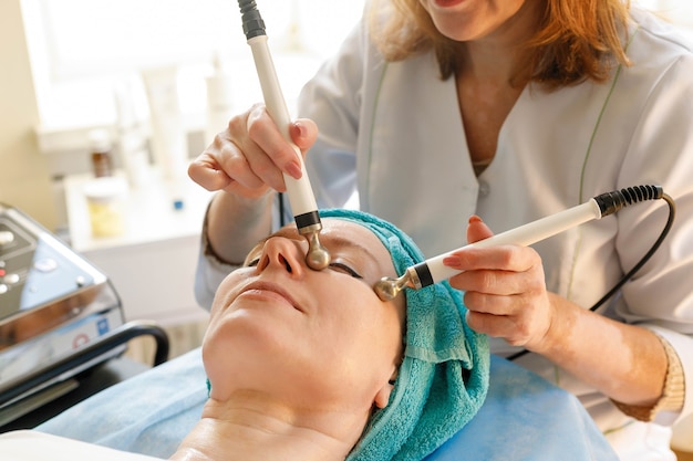 Koncepcja ludzie, uroda, spa, kosmetologia i technologia - Kosmetolog wykonuje zabieg mikroprądowej terapii skóry twarzy pięknej, młodej kobiety w gabinecie kosmetycznym