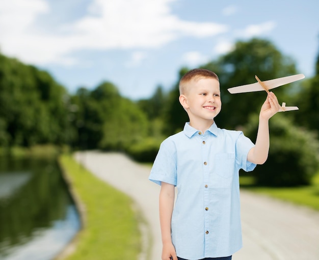 Zdjęcie koncepcja marzeń, przyszłości, hobby, natury i dzieciństwa - uśmiechnięty mały chłopiec trzymający w dłoni drewniany model samolotu nad parkowym tłem