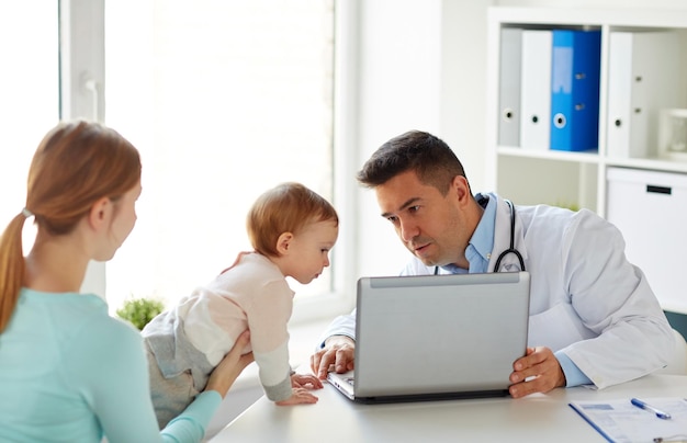 koncepcja medycyny, opieki zdrowotnej, pediatrii i ludzi - szczęśliwa kobieta z dzieckiem i lekarz z laptopem w klinice