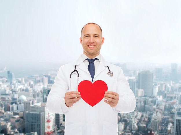 koncepcja medycyny, zawodu, miłości i opieki zdrowotnej - uśmiechnięty mężczyzna lekarz z czerwonym sercem i stetoskopem na tle miasta