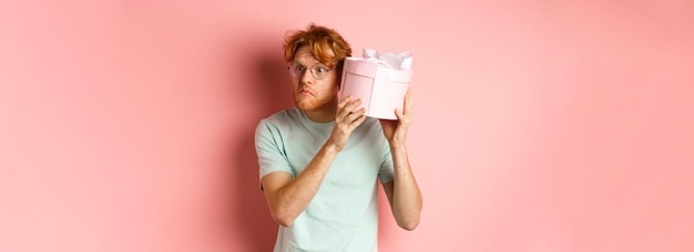 Koncepcja miłości i wakacji zaintrygowana rudowłosy facet przyciska ucho do pudełka i potrząsa prezentem, zgadując, co insi