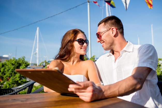 koncepcja miłości, randek, ludzi i jedzenia - uśmiechnięta para w okularach przeciwsłonecznych, trzymająca menu i patrząca na siebie w kawiarni
