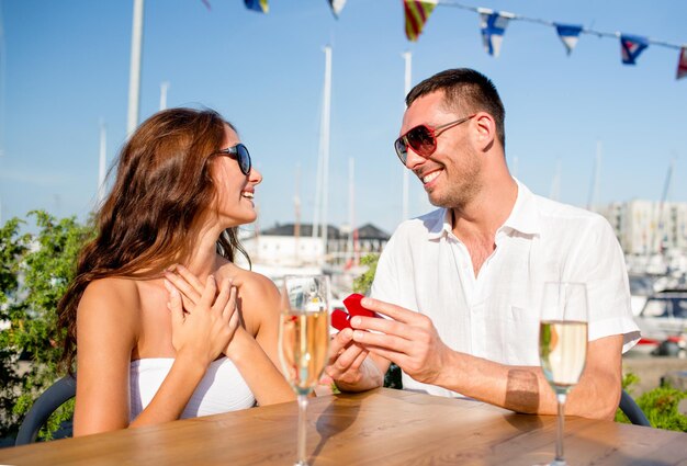 koncepcja miłości, randek, ludzi i wakacji - uśmiechnięta para w okularach przeciwsłonecznych z szampanem i małym czerwonym pudełku patrzącym na siebie w kawiarni