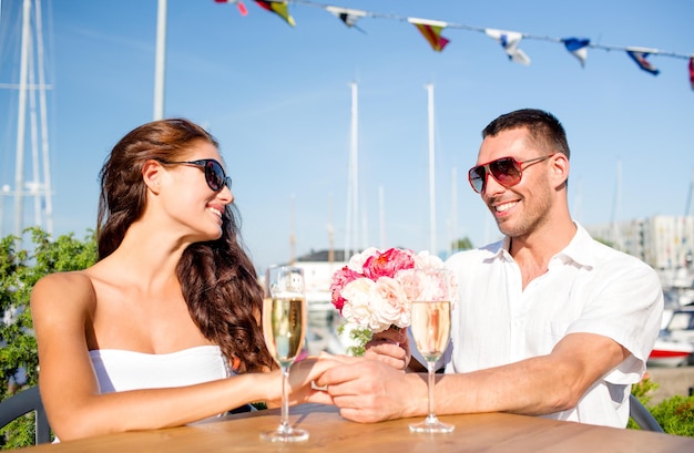 koncepcja miłości, randki, szczęścia i ludzi - uśmiechnięta para w okularach przeciwsłonecznych z bukietem kwiatów i kieliszkami szampana, patrząca na siebie w kawiarni