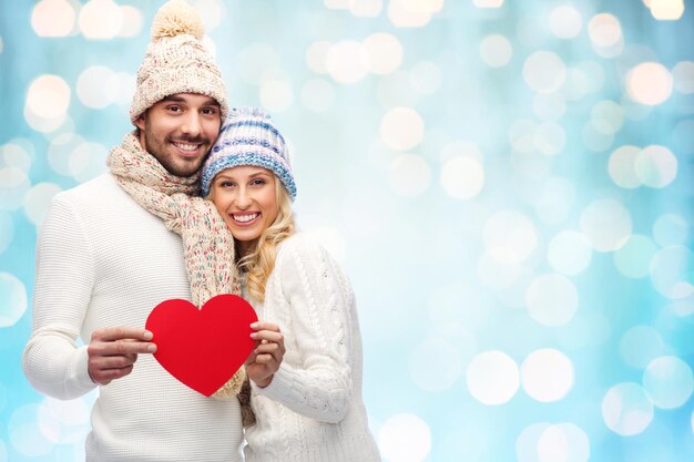 koncepcja miłości, walentynek, pary, bożego narodzenia i ludzi - uśmiechnięty mężczyzna i kobieta w zimowych czapkach i szaliku trzymających czerwony papierowy kształt serca na niebieskim tle świateł świąt