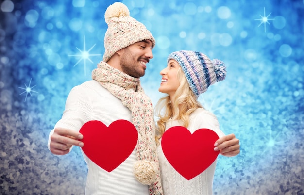 koncepcja miłości, walentynki, pary, bożego narodzenia i ludzi - uśmiechnięty mężczyzna i kobieta w zimowych czapkach i szaliku trzymających czerwone papierowe kształty serca na niebieskim tle brokatu i świątecznych świateł