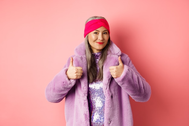 Koncepcja mody i zakupów. Azjatycka modna dama w płaszczu zimowym pokazująca kciuk w górę, uśmiechnięta i zadowolona, polecająca promocję firmy, różowe tło.
