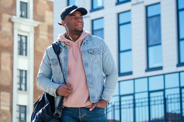 Koncepcja mody ulicznej - stylowy przystojny afroamerykanin w różowej koszuli, dżinsowej kurtce i czarnej czapce na tle okien centrum biznesowego