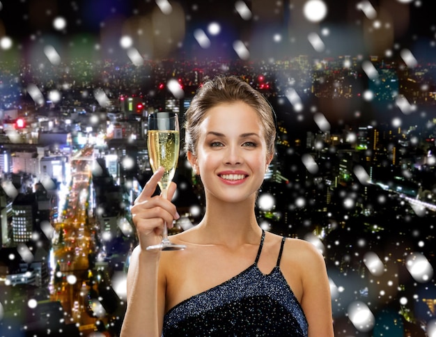 koncepcja napojów, świąt, świąt, ludzi i uroczystości - uśmiechnięta kobieta w stroju wieczorowym z lampką wina musującego na tle zaśnieżonego miasta
