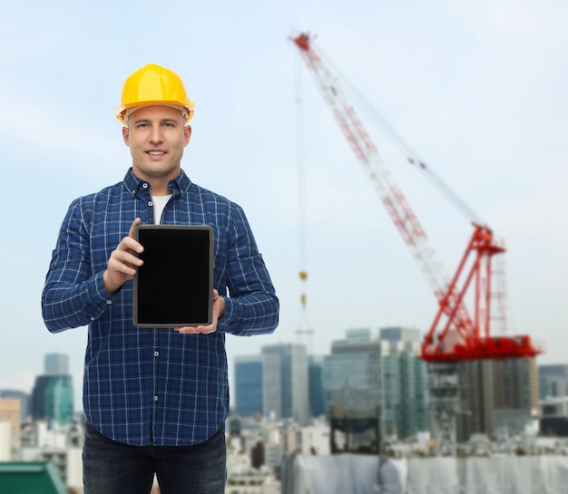 koncepcja naprawy, budowy, ludzi i konserwacji - uśmiechnięty mężczyzna budowniczy lub pracownik fizyczny w kasku pokazujący pusty ekran komputera typu tablet pc na tle placu budowy miasta