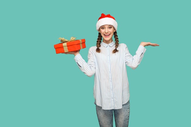Koncepcja nowego roku. szczęśliwa piękna dziewczyna w jasnoniebieskiej koszuli w paski i czerwonej czapce świątecznej, trzymająca czerwone pudełko i patrząca w kamerę z zaskoczoną miną. kryty na białym tle na zielonym tle.