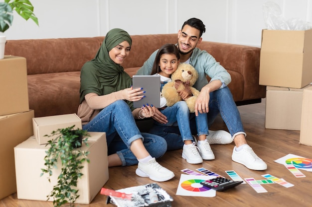 Koncepcja Nowoczesnej Komunikacji Zdalnej. Portret Pozytywnej Arabskiej Rodziny Wykonującej Wirtualną Rozmowę Wideo Z Przyjaciółmi Lub Dziadkami Za Pomocą Tabletu, Siedzącej Na Dywanie Podłogowym Z Farbą, Rozmawiającej Online