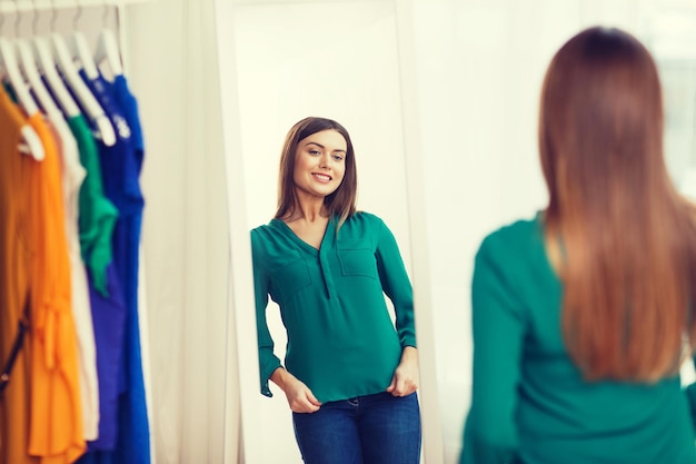 koncepcja odzieży, mody, stylu i ludzi - szczęśliwa kobieta wybierająca ubrania i pozująca przed lustrem w domowej garderobie