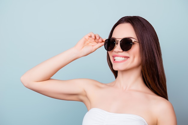 Koncepcja okularów przeciwsłonecznych. Młoda śliczna brunetka z zębami w letnim stroju i modnych okularach przeciwsłonecznych stoi na jasnoniebieskiej przestrzeni, poprawiając swoje okulary. Jest taka stylowa i atrakcyjna
