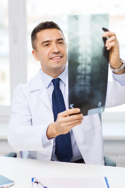 koncepcja opieki zdrowotnej, rontgen, ludzi i medycyny - uśmiechnięty mężczyzna lekarz w białym fartuchu patrzący na zdjęcie rentgenowskie w gabinecie lekarskim