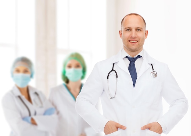 koncepcja opieki zdrowotnej, zawód, praca zespołowa, ludzie i medycyna - uśmiechnięty mężczyzna lekarz ze stetoskopem w białym fartuchu nad grupą medyków