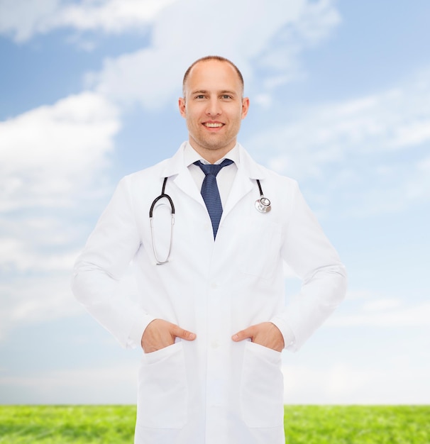 koncepcja opieki zdrowotnej, zawodu, środowiska i medycyny - uśmiechnięty mężczyzna lekarz ze stetoskopem w białym fartuchu na tle przyrody