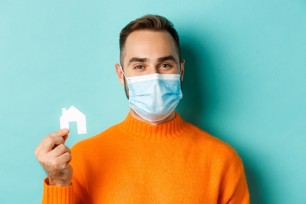 Koncepcja pandemii nieruchomości i koronawirusa. Zbliżenie: dorosły mężczyzna w masce medycznej, trzymając mały papierowy dom maket i uśmiechnięty, szukając mieszkania, jasnoniebieskie tło.