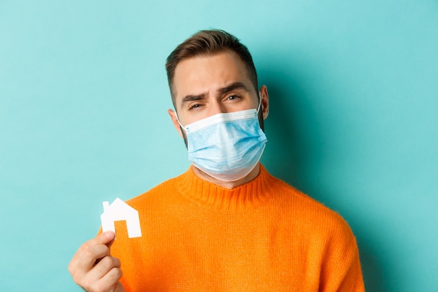 Koncepcja pandemii nieruchomości i koronawirusa. Zbliżenie: smutny mężczyzna w masce na twarz, pokazując maskę i krzywiąc się zdenerwowany, stojąc na jasnoniebieskim tle.