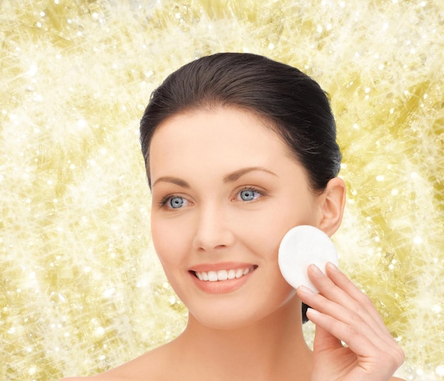 koncepcja piękna, ludzi i zdrowia - piękna uśmiechnięta kobieta czyści skórę twarzy za pomocą wacika na żółtym tle światła