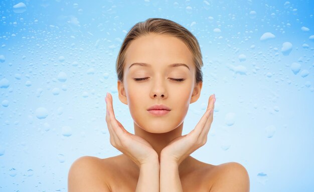 Zdjęcie koncepcja piękna, ludzi, pielęgnacji skóry i zdrowia - twarz młodej kobiety i ręce nad kroplami wody na niebieskim tle