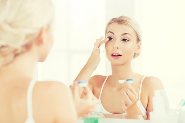 Zdjęcie koncepcja piękna, wzroku, wzroku, okulistyki i ludzi - młoda kobieta zakładająca soczewki kontaktowe w lustrze w domowej łazience