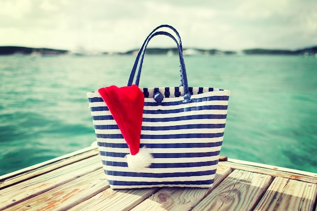 Zdjęcie koncepcja plaży, lata, wakacji, świąt i akcesoriów - zbliżenie torby plażowej i kapelusza pomocnika świętego mikołaja na drewnianym molo