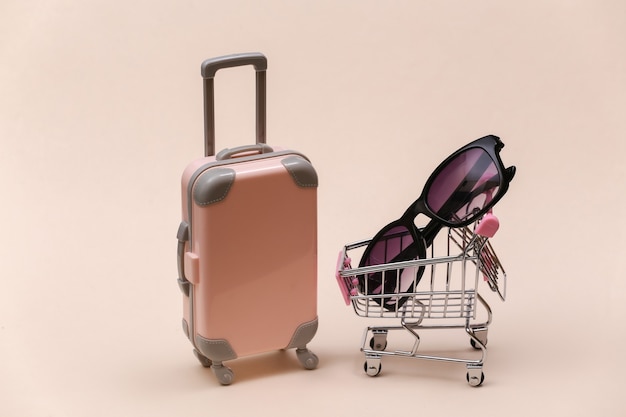Zdjęcie koncepcja podróży i zakupów. mini walizka podróżna z tworzywa sztucznego i wózek do supermarketu z okularami przeciwsłonecznymi na beżowym tle.