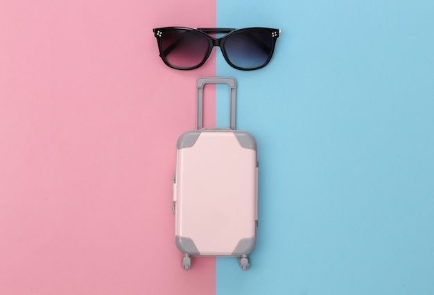 Koncepcja podróży. Mini walizka podróżna z tworzywa sztucznego i okulary przeciwsłoneczne na różowym niebieskim tle pastelowych. Minimalistyczny styl. Widok z góry, układ płaski