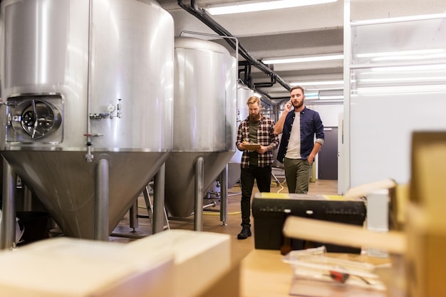 Zdjęcie koncepcja produkcji, biznesu i ludzi - mężczyźni ze schowkiem pracujący w browarze rzemieślniczym lub zakładzie piwa i dzwoniący na smartfonie