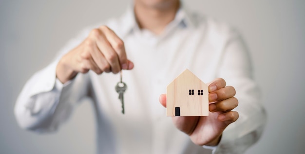 Koncepcja przeprowadzki do nowego domu, Dłoń trzymająca klucz do drewnianego domu i domu, Inwestycja w nieruchomość i hipoteka finansowa nieruchomości, Pośrednicy w obrocie nieruchomościami trzymają klucze do drewnianego domu