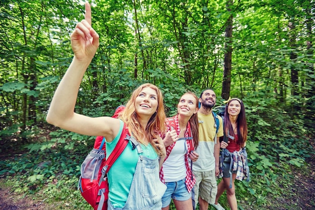 koncepcja przygód, podróży, turystyki, wędrówek i ludzi - grupa uśmiechniętych przyjaciół z plecakami wskazującymi palcem w górę w lesie