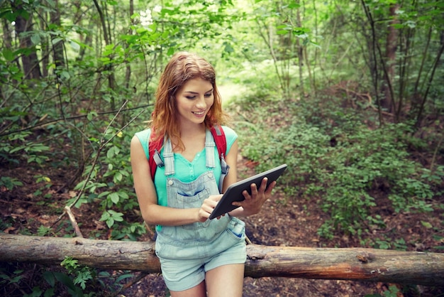 koncepcja przygód, podróży, turystyki, wędrówek i ludzi - szczęśliwa młoda kobieta z plecakiem i komputerem typu tablet, siedząca na pniu zwalonego drzewa w lesie