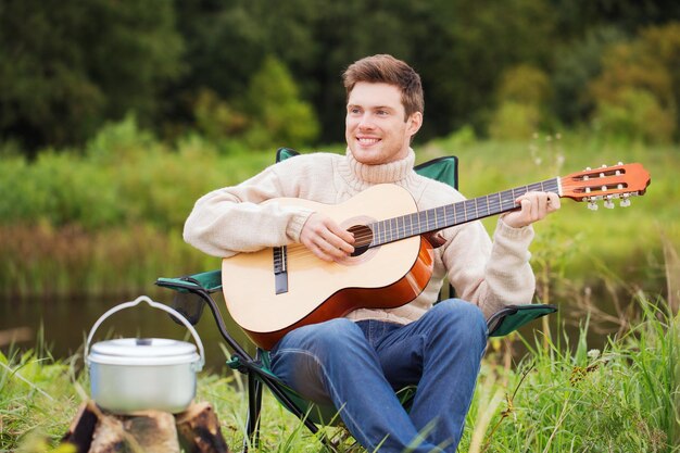 koncepcja przygody, podróży, turystyki i ludzi - uśmiechnięty mężczyzna z gitarą i gotowanie jedzenia na ognisku na kempingu