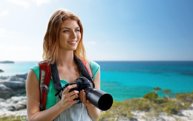 koncepcja przygody, podróży, turystyki, wędrówek i ludzi - szczęśliwa młoda kobieta z plecakiem i aparatem fotografującym nad brzegiem morza lub plażą