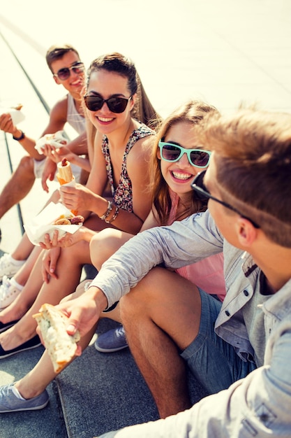 koncepcja przyjaźni, wypoczynku, lata i ludzi - grupa uśmiechniętych przyjaciół w okularach przeciwsłonecznych siedzących z jedzeniem na placu miejskim
