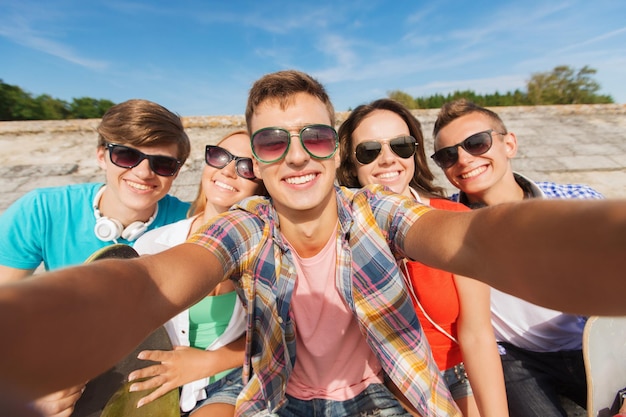 koncepcja przyjaźni, wypoczynku, lata, technologii i ludzi - grupa uśmiechniętych przyjaciół z deskorolką robi selfie na świeżym powietrzu