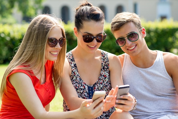 koncepcja przyjaźni, wypoczynku, lata, technologii i ludzi - grupa uśmiechniętych przyjaciół ze smartfonem siedzących na trawie w parku