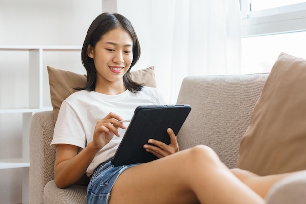 Koncepcja przytulnego stylu życia Młoda kobieta leżąca na kanapie i używająca tabletu do surfowania po mediach społecznościowych w domu