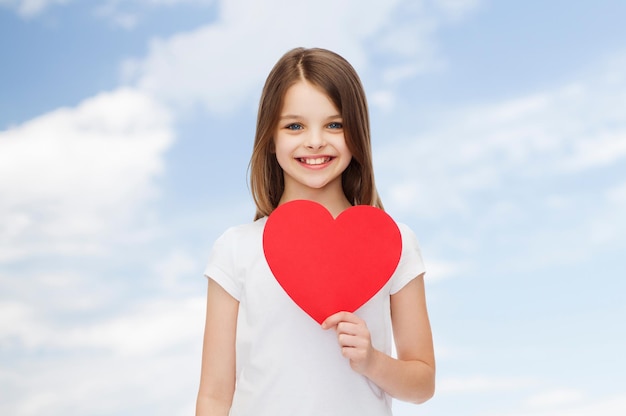 koncepcja reklamy, dzieciństwa, lata, miłości i ludzi - uśmiechnięta dziewczyna w białej koszulce trzymająca czerwone serce na naturalnym tle